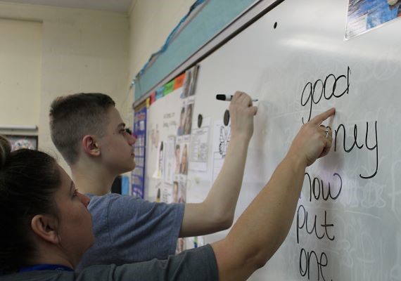 Student writing on whiteboard with teacher standing beside him