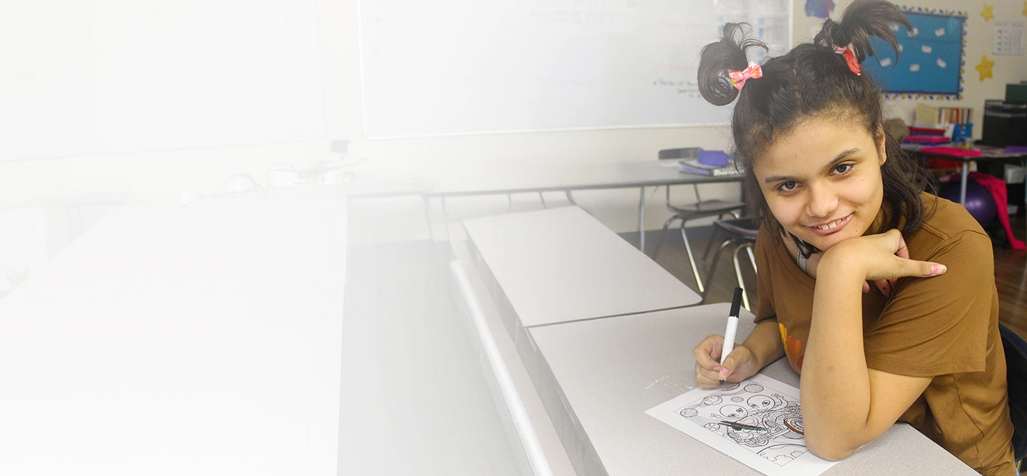 Student sitting at a classroom desk, smiling while drawing on a worksheet