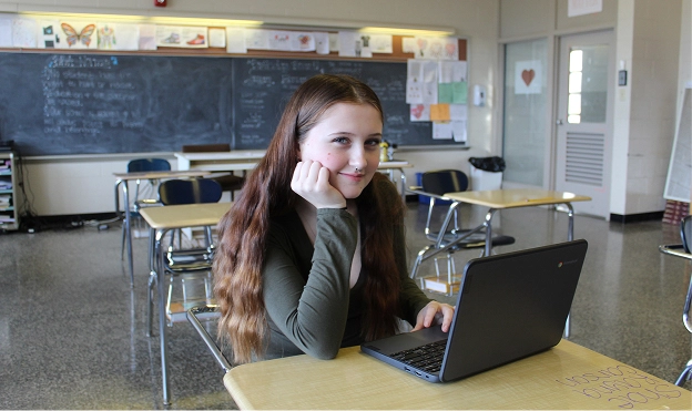 Student working on a laptop at a desk in a classroom with desks and a chalkboard in the background.