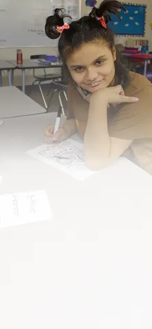 Student sitting at a classroom desk, smiling while drawing on a worksheet